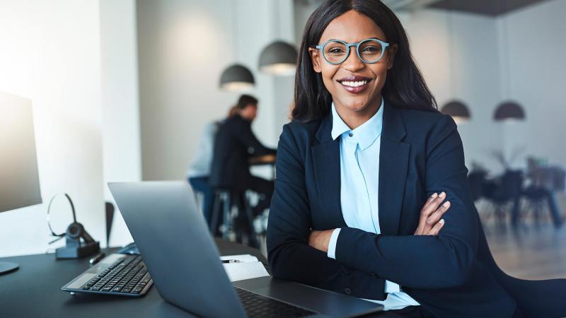 eine Geschäftsfrau afrikanischer Abstammung sitzt an einem Schreibtisch und schaut lächelnd in die Kamera. Sie trägt eine Brille und lächelt, während sie in einem Büro eines Schifffahrtsbetriebs am Schreibtisch sitzt.