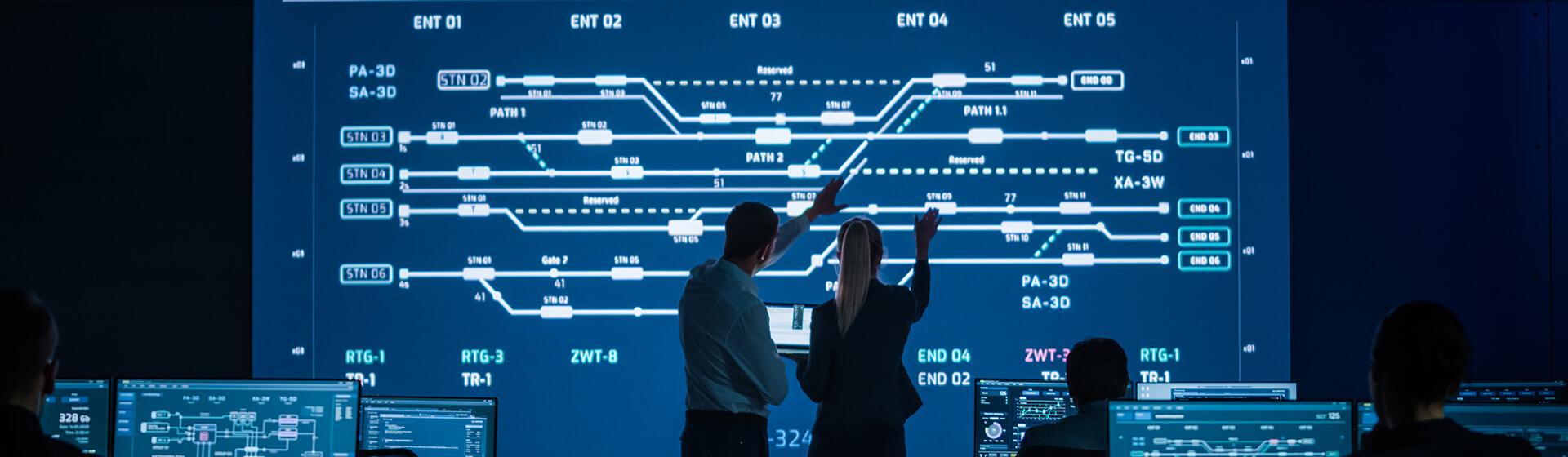 en profesjonell mann og kvinne i et kontrollrom med en stor skjerm som viser jernbanespor på en maritim shipping terminal.