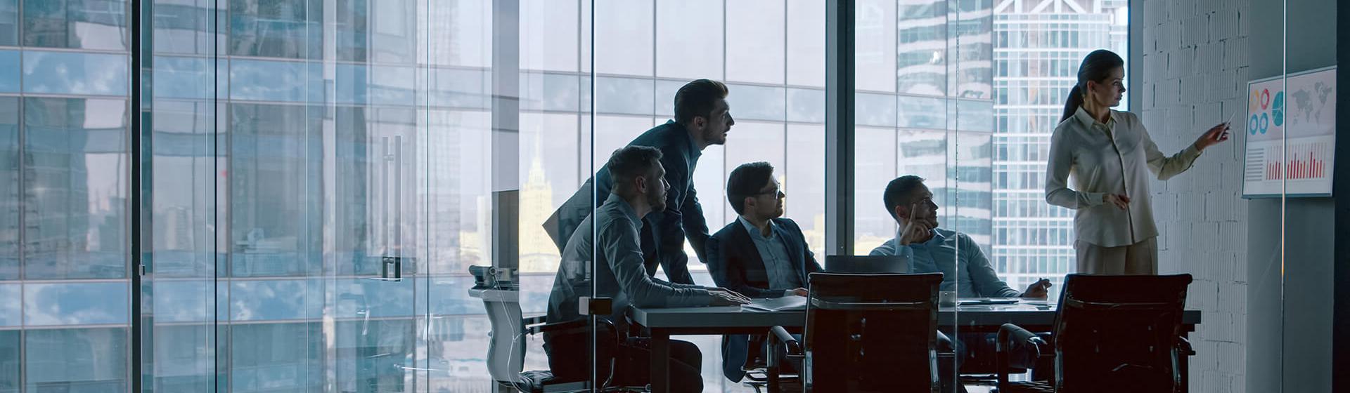 Une salle de bureau en verre, avec une employée montrant des données et des graphiques de transport maritime sur un écran accroché au mur, avec trois hommes assis à table qui regardent l’écran. Un quatrième homme se tient debout et regarde également l’écran.