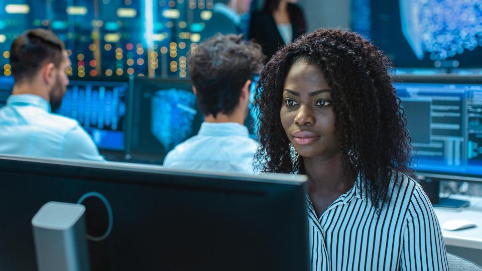 eine Frau afrikanischer Abstammung sitzt vor einem Computerbildschirm. Weitere Mitarbeitende sitzen an eigenen Computern im Hintergrund.
