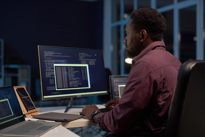 رجل متخصص من أصل أفريقي يجلس أمام لوحة مفاتيح الكمبيوتر في مكتبه. شاشة الكمبيوتر محاطة بهاتف ذكي وجهاز لوحي رقمي في علبته وجهاز كمبيوتر محمول.